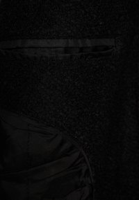 Svart ullkappa närbild, med en texturerad tyg, slät foder och en dold fickdetalj. Interiören är av svart satinliknande material.