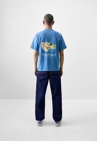 Šviesiai mėlyna medvilninė marškinėliai su apvaliu piešiniu, kurį puošia kalnai, o ant nugaros yra užrašai "Penfield" ir "Est 1975", derinami su tamsiai mėlynomis džinsais.