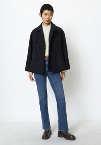 Svart dobbeltspent frakk med bred krage over en lys grønn skjorte, sammen med blå rette jeans og mørkebrune kraftige sko.