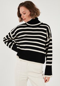 Mustmustriibuline must-valge turtleneck-sviiter ribakudumist, millel on lühendatud disain ja laiad varrukad kontrastse äärisega.
