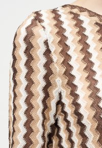 Κοντινό πλάνο πλεκτού υφάσματος με κυματιστές ζιγκ-ζαγκ ρίγες σε καφέ, μπεζ και λευκούς τόνους στον ώμο ενός ενδύματος.