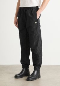 Schwarze Jogginghosen mit elastischem Bund, seitlichen Taschen, strukturiertem Finish und kleinem grünen Logo. Kombiniert mit schwarzen Knöchelstiefeln.