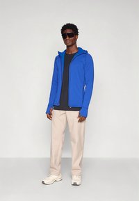 Kék cipzáras kapucnis kabát, alatta fekete póló, világosbézs nadrág, és szürke sportcipő. A modell napszemüveget visel.