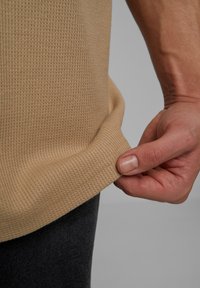 Bežová textúrovaná tkanina s vaflovým vzorom, ruky drží okraj, v kombinácii s tmavosivým materiálom. Detail ležérneho oblečenia.
