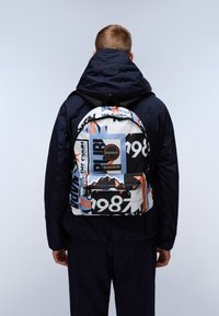 Le sac à dos présente un mélange de motifs géométriques colorés et de texte sur fond blanc, avec des bretelles noires et une poche zippée à l'avant.