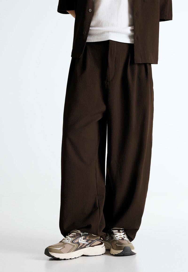 Személy laza, sötétbarna nadrágot, fehér inget, sötétbarna zakót és bézs, fehér és fekete sportcipőt visel, világos háttér előtt áll.