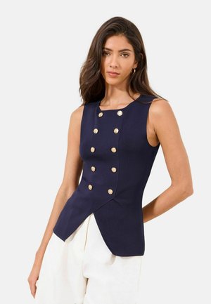 Naine, kes kannab varrukateta tumesinist kahekordse nööpimisega topi kuldsete nööpidega ja valgeid pükse, seisab ühevärvilise tausta ees.