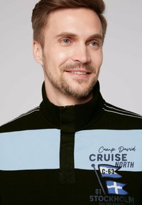Pullover di cotone nero con colletto alto, caratterizzato da strisce orizzontali blu, grafiche ricamate e cuciture di accento bianche.