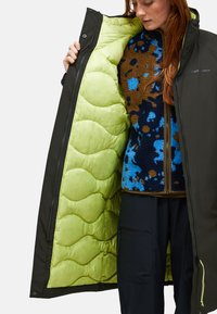 Person mit roten Haaren zeigt das leuchtend grüne, gesteppte Futter einer schwarzen Zip-Jacke, trägt ein blaues und braunes Fleece mit Muster und schwarze Hosen.