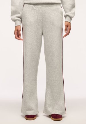 Pantaloni della tuta grigio chiaro a gamba larga con strisce laterali color granata indossati con scarpe granata, mostrando dalla vita ai piedi con le mani ai lati.
