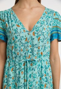 Lyseblå kjole med blomstermønster, korte ærmer, V-hals, knapper ned langs fronten og et bånd i taljen. Har orange og teal accenter.