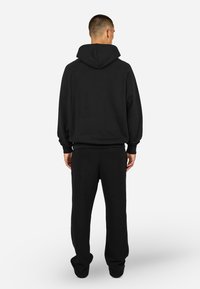 Μαύρο φούτερ με κουκούλα και ραγλανιστά μανίκια, συνδυασμένο με μαύρο υφασμάτινο παντελόνι. Και τα δύο κομμάτια έχουν χαλαρή εφαρμογή και ρίγες στα μανίκια.