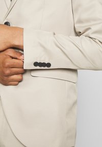 Giacca beige con una texture morbida. Presenta bottoni neri sui polsini e una tasca sul petto. Vestibilità sartoriale con spalle strutturate.