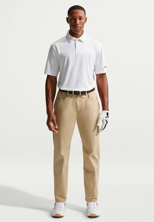 Homme debout portant un polo blanc à manches courtes, un pantalon beige, un gant de golf blanc à la main droite, une ceinture noire et des baskets blanches sur fond blanc.