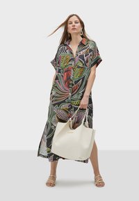 Abito a maniche corte con una base nera e motivi a foglia multicolori. Il modello tiene una borsa tote bianca, indossando sandali dorati con cinturini.