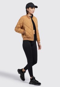 Gevilte leren bomberjack in het bruin met geribbelde manchetten, gecombineerd met zwarte leggings en zwarte sneakers. De pet is zwart, wat de sportieve look compleet maakt.