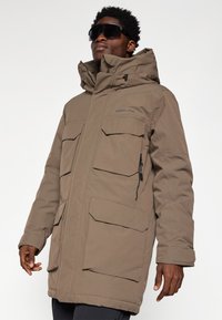 Brun isoleret parka med høj krave, flere frontlommer og justerbare manchetter. Har lynlåslukning og hætte. Glat tekstur.