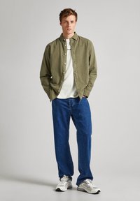 Olijfgroene button-up shirt over een wit T-shirt, gecombineerd met blauwe jeans en grijze sneakers. Het shirt heeft een relaxed fit en een kraag.