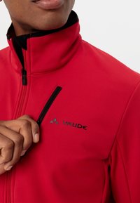 Veste softshell rouge avec un col montant, une fermeture éclair noire, et une petite poche poitrine zippée. Présente un logo noir sur le tissu.