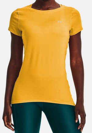 Vrouw die een geel sportshirt met korte mouwen en donkergroene leggings draagt, staand tegen een effen achtergrond.