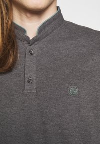 Polo gris foncé avec un col montant, deux boutons noirs, et un logo brodé vert turquoise sur le côté gauche. Texture douce.