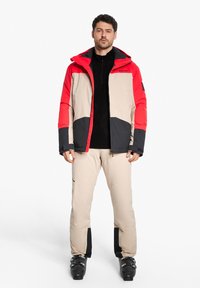Herren-Skijacke in rot, beige und schwarz mit Kapuze, kombiniert mit beigen Skihosen. Beide haben eine glatte Textur und ein ergonomisches Design.