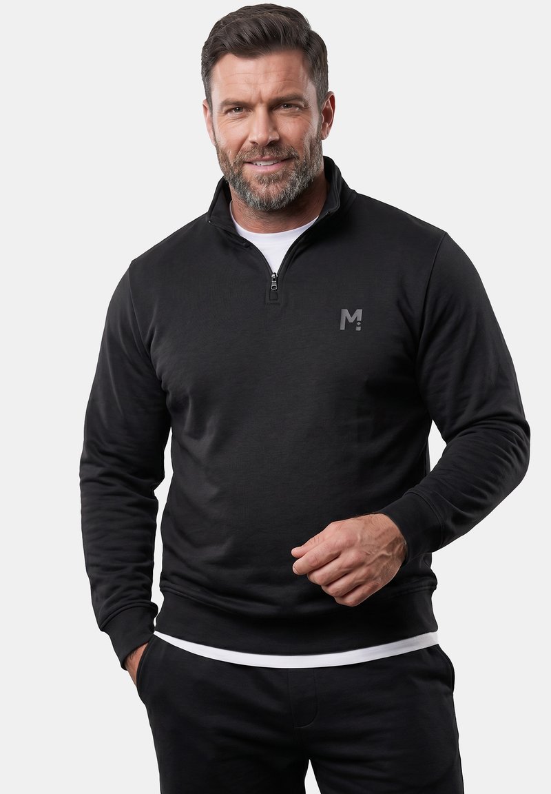 Man met kort haar en baard draagt een zwarte quarter-zip trui met een klein 'M' logo, een wit ondershirt en donkere broek, staat en glimlacht.
