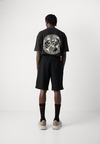 En person står med ryggen til iført en svart oversized T-skjorte med hvit boksing grafikk, svarte shorts, svarte sokker med rødt tekst, og beige sneakers.