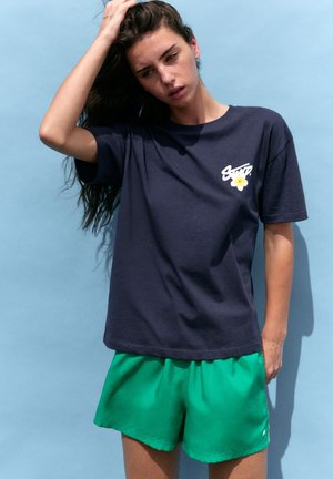 Femme aux longs cheveux foncés portant un t-shirt bleu marine avec un logo et un short vert, debout devant un fond bleu clair.