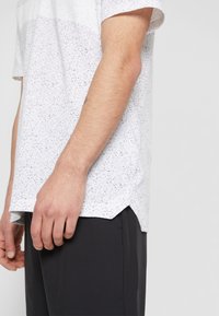 T-shirt blanc moucheté avec un col rond, des manches courtes et un ourlet avec des fentes latérales, associé à un pantalon noir. Tissu texturé.