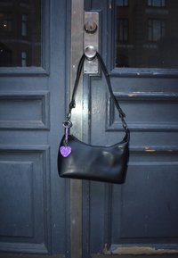 Czarna syntetyczna torebka o zakrzywionym kształcie, wyposażona w pasek oraz brelok w kształcie fioletowego migoczącego serca. Umieszczona na tle niebieskich drzwi.