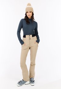 Vrouw in beige skibroek, grijze langarmtop, beige gebreide muts en grijze skischoenen, staande met handen in de zakken op een witte achtergrond.