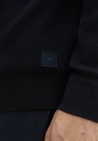 Tamsus megztinis su grioveliu apatinėje dalyje ir rankogaliais, su mažu juodu stačiakampiu logotipo žymekliu apačioje. Lygi tekstūra, prigludusi forma.