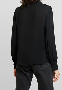 Blusa nera a maniche lunghe con una trama liscia, orlo arrotondato e polsini con bottoni. Presenta maniche plissettate per un tocco di originalità.