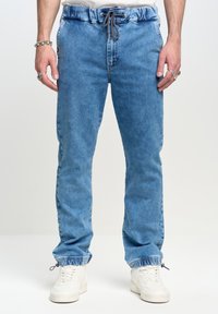 Heledad sinine teksane jogger püksid elastse vöökohtaga, paeladetaili ja elastsete mansettidega. Koos valgete tossudega.