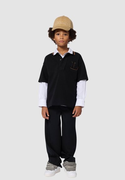 Czarna koszulka polo z krótkimi rękawami i kieszonką na piersi, noszona na białej koszuli. Czarne szerokie spodnie oraz beżowa czapka z wytłoczonym napisem.