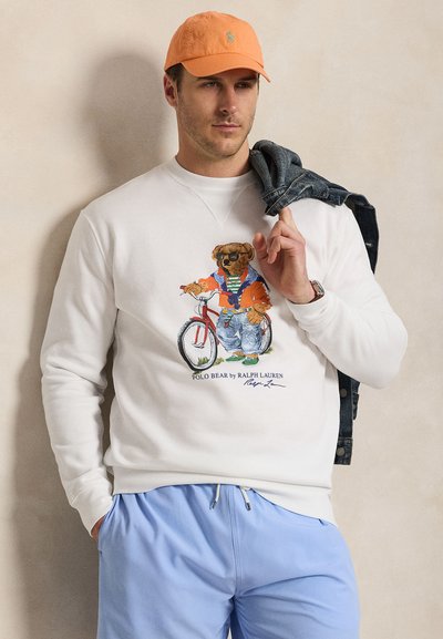 Férfi narancssárga sapkával, fehér pulóverrel medvés biciklis grafikával, világoskék nadrágban, farmerdzsekit tart a vállán.