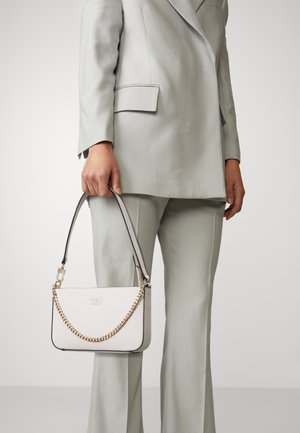 Personne en costume gris clair tenant un petit sac à main blanc avec une chaîne dorée et le logo "GUESS" à l'avant sur un fond uni.