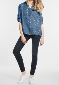 Denimskjorta i mörkblått med vitt fågeltryck, avslappnad passform, uppvikta ärmar; kombinerad med mörka skinnyjeans och vita sneakers.