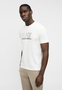 Λευκό βαμβακερό μπλουζάκι με μαύρο λογότυπο που φέρει το κείμενο "EA7" και "EMPORIO ARMANI". Κοντά μανίκια και στρογγυλή λαιμόκοψη, εφαρμοστό στυλ.