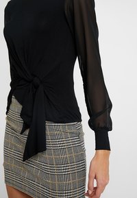 Black top with knotted front detail and sheer long sleeves, paired with a fitted check-patterned skirt in black, white, and gold accents.