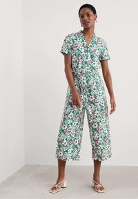 Sieviete stāv floralā īsās piedurknes kombinezonā ar platām cropped biksēm un baltām siksnām blokveida papēžos, neitrālā fonā.