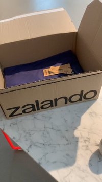 Kartonska škatla z natisnjenim "zalando" v črni barvi. Vsebuje zloženo modro oblačilo in etiket. Škatla je odprta na marmorni površini.