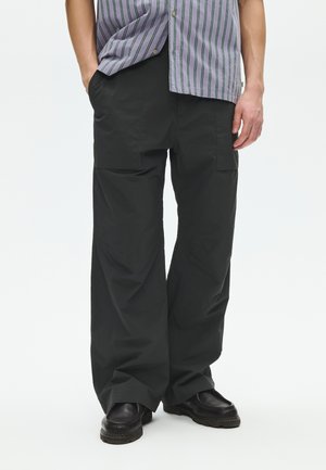 Άνδρας που φοράει μαύρο παντελόνι cargo με φαρδιά μπατζάκια, μαύρα παπούτσια και ριγέ κοντομάνικο πουκάμισο, μερικώς μέσα από το παντελόνι, στέκεται με τα χέρια στις τσέπες.