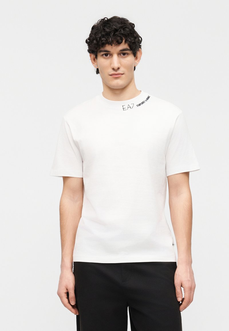 Νεαρός άνδρας με κοντά σγουρά μαλλιά, ντυμένος με λευκή μπλούζα με το κείμενο "EA7 Emporio Armani" κοντά στον λαιμό και μαύρο παντελόνι.