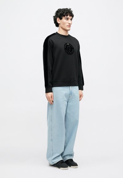 Melnā džemperis ar apaļu kakla izgriezumu un teksturētu logotipa detaļu, kopā ar gaiši zilām platām džinsa biksēm un melniem apaviem.