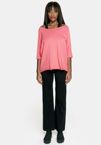Luźna koralowo-różowa bluzka o szerokim dekolcie i rękawach do łokcia, zestawiona z czarnymi, rozszerzanymi spodniami i czarnymi butami sportowymi.
