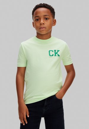 Jongen met kort krullend haar, gekleed in een lichtgroen T-shirt met CK-logo en donkere jeans, staand met één hand in zijn zak tegen een grijze achtergrond.