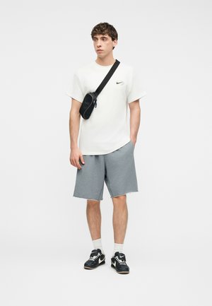 Νέος άνδρας που φοράει λευκό t-shirt Nike, γκρι σορτς, μαύρη τσάντα χιαστί, λευκές κάλτσες και μαύρα αθλητικά παπούτσια Nike, στέκεται μπροστά σε λευκό φόντο.