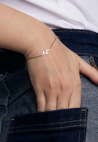 Bransoletka w kolorze srebra z delikatnym wzorem liścia, połączona cienkim łańcuszkiem, noszona na ręce opierającej się na dżinsach.